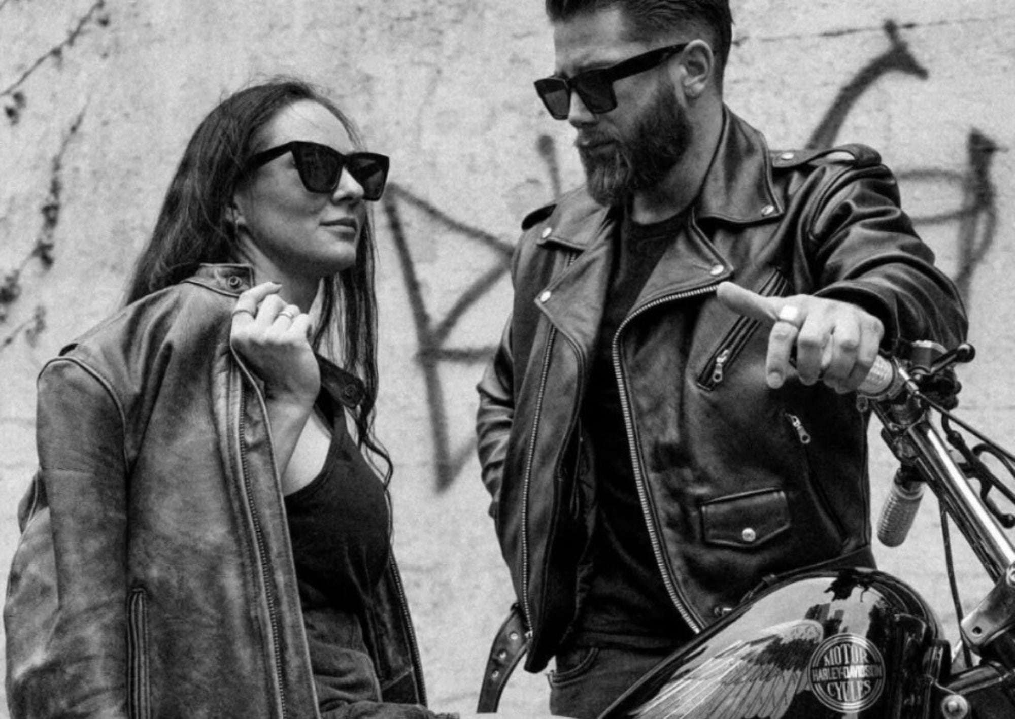 Man and woman in Beck Northeaster Flying Togs leather motorcycle jackets on a motorcycle with a graffiti-covered wall in the background
